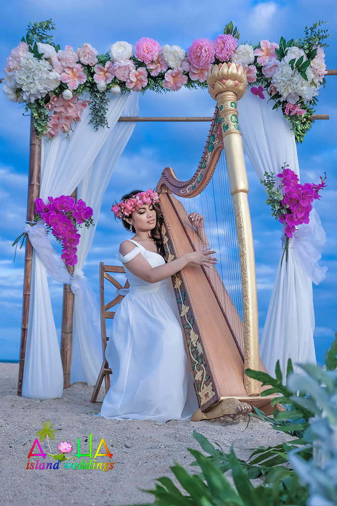 Harpist in a white dress and pink flower crown playing a golden harp under a beach arch of pink peonies, white hydrangeas and orchids